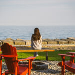 Woman in a a sweater and jeans, with long brown hair sits on wooden bench by firepit looking at Lake Erie