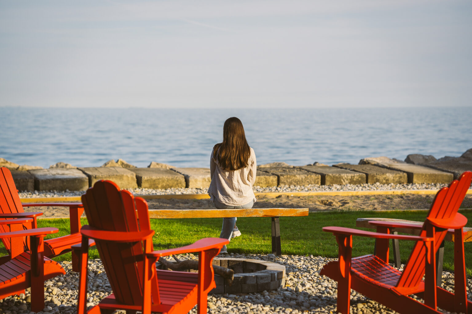 Woman in a a sweater and jeans, with long brown hair sits on wooden bench by firepit looking at Lake Erie