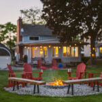 Evening at a cottage on Lake Erie, Kingsville, with sauna and lit-up cottage in background and fire pit, red muskoka chairs, and benches in the foreground