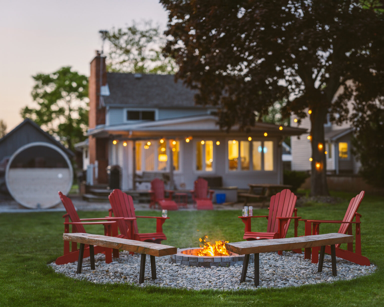 Evening at a cottage on Lake Erie, Kingsville, with sauna and lit-up cottage in background and fire pit, red muskoka chairs, and benches in the foreground