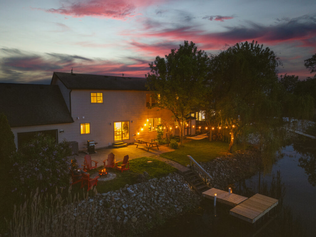 Night view with string lights of back of two-story home on Cedar Creek, with a dock, red muskoka chairs, hammock, picnic table.