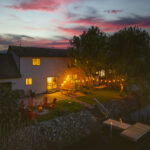 Sunset view of two story vacation rental with string lights on back patio, dock, fire in the fire pit, and red chairs. (Ironwood Cottages on Lake Erie in Kingsville)